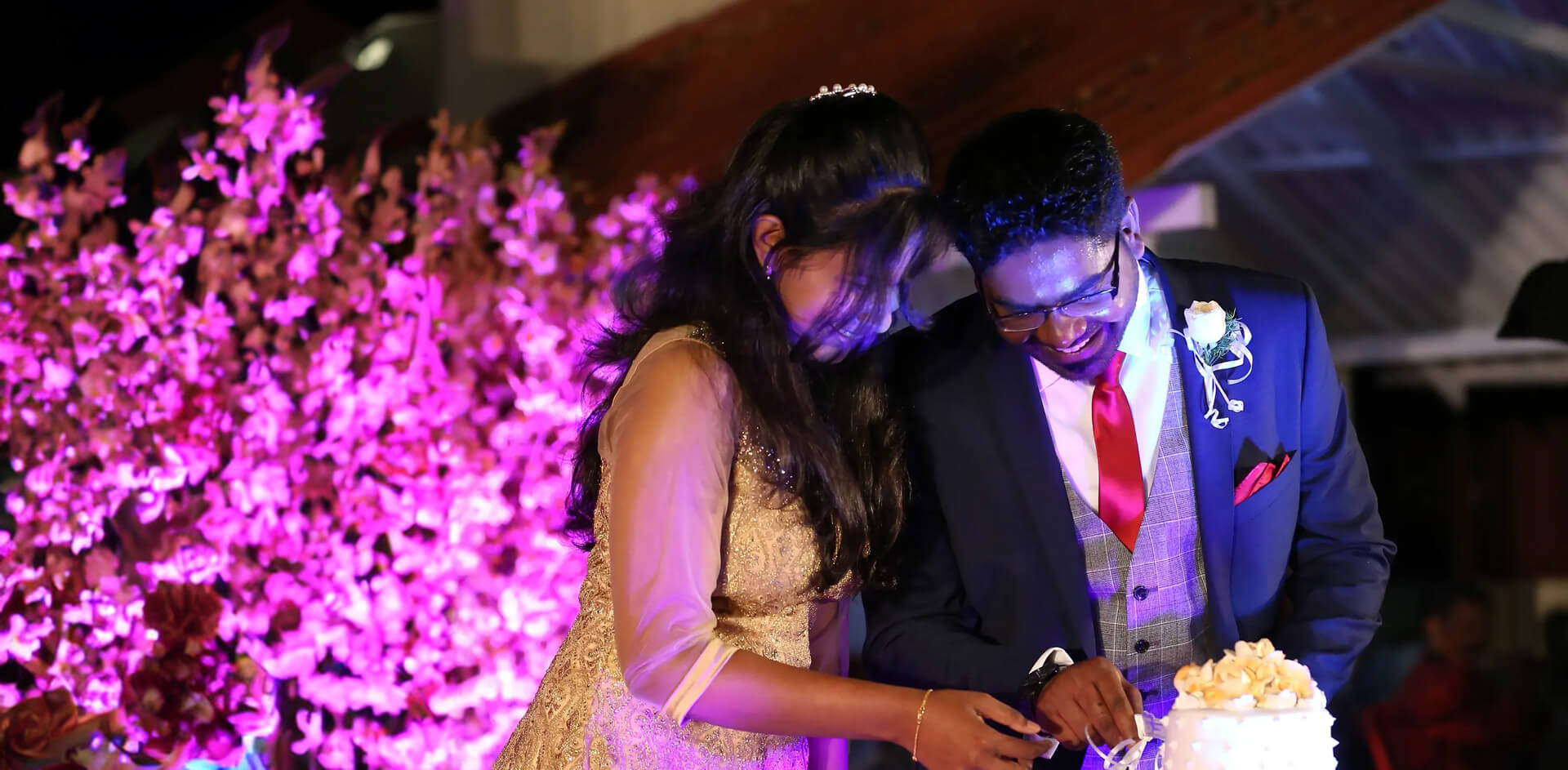 Bride and groom cutting their wedding cake under pink floral lighting at a reception