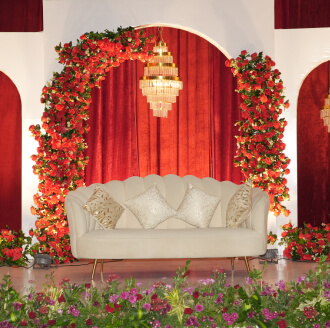 Wedding stage décor with red drapes, a floral arch and a white sofa by our wedding event management company
