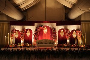 Reception stage decoration with red and gold floral arches, chandeliers and candle stands
